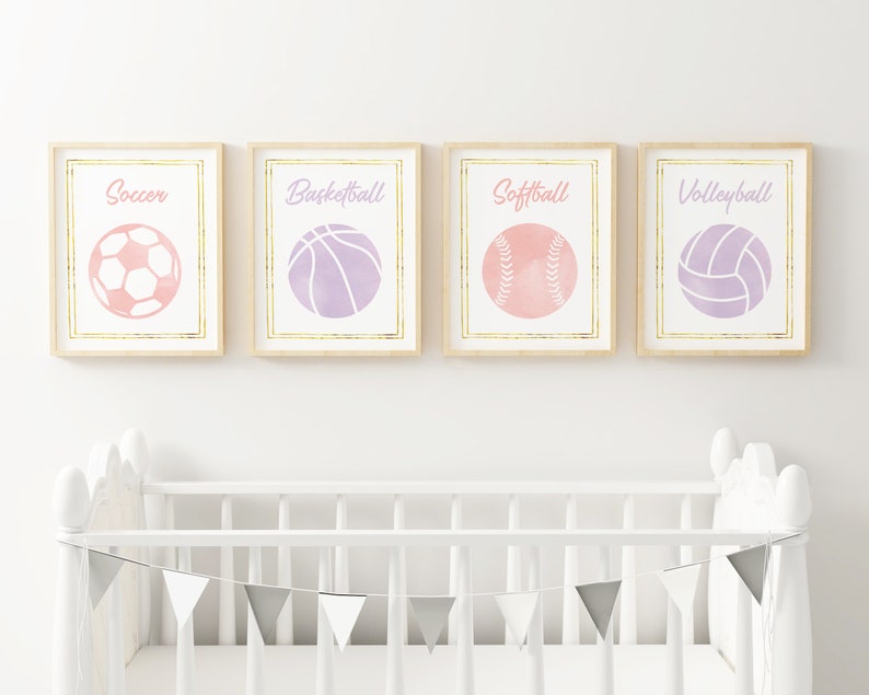May include: Four framed prints with watercolor illustrations of sports balls. Each print features a different sport: soccer, basketball, softball, and volleyball. The balls are pink and purple with gold outlines. The prints are displayed on a white wall above a white crib.