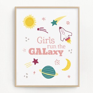 May include: A framed print with a gold border featuring a space-themed design. The artwork includes a yellow sun, stars, a rocket ship, a planet, and a crescent moon. The text "Girls run the GALAXY" is displayed in pink and coral lettering.