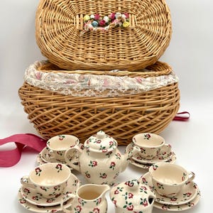May include: A tea set featuring a teapot, creamer, sugar bowl, and four cups with saucers, all decorated with a red and green floral pattern on a cream background. The set is displayed in front of a woven wicker picnic basket with a floral lid.
