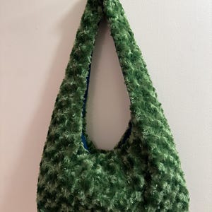 May include: A green, fluffy shoulder bag with a soft, textured exterior. The bag has a rounded shape and a blue interior lining. The strap is integrated into the bag's design, creating a seamless look. The bag is hanging against a white wall.