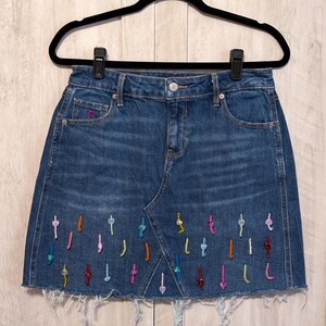 May include: A dark blue denim mini skirt with a frayed hem. The skirt is decorated with colorful beaded embellishments in the shape of small arrows.