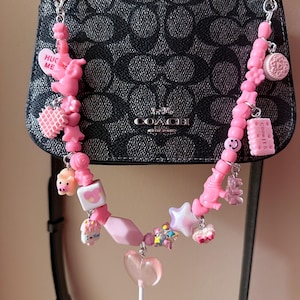 May include: A pink charm bracelet with various charms, including a heart-shaped lollipop, a star, a pig, a waffle, a cookie, and a charm that says "Hug Me". The bracelet is attached to a black Coach purse.