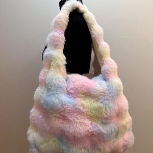 May include: A soft, plush shoulder bag featuring a multi-colored faux fur exterior with a pastel tie-dye pattern in shades of pink, blue, yellow, and white. The bag has a wide, integrated strap and a slouchy, unstructured rectangular shape.