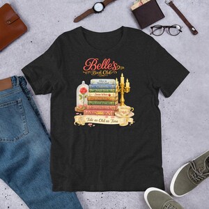 May include: Dark gray t-shirt featuring a graphic of a stack of books with titles like "Alice in Wonderland" and "Snow White." The design includes a rose in a glass dome, a candelabra, and the text "Belle's Book Club."