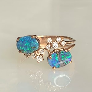 May include: A rose gold ring featuring two oval-cut opals with vibrant blue and green iridescence. Small, clear gemstones accent the opals. The ring has a split shank design.