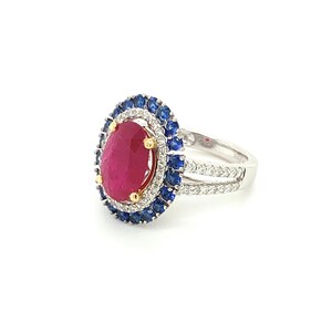 Op de afbeelding: Een zilveren ring met een grote ovale robijn omringd door een halo van kleinere diamanten en saffieren.