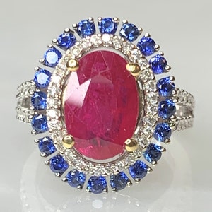 Op de afbeelding: Een ring met een grote ovale robijn, omringd door een halo van kleinere diamanten en blauwe saffieren.