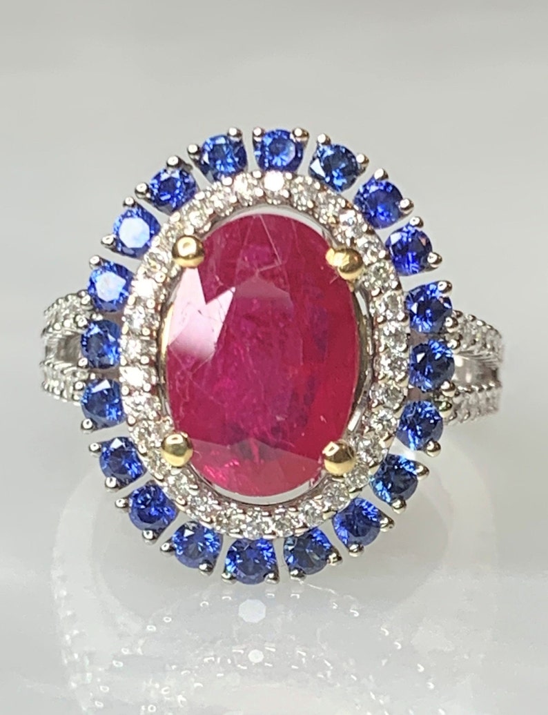Op de afbeelding: Een ring met een grote ovale robijn, omringd door een halo van kleinere diamanten en blauwe saffieren.
