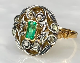 May include: A gold and silver ring featuring an emerald gemstone surrounded by smaller diamonds. The ring has a delicate, floral design.