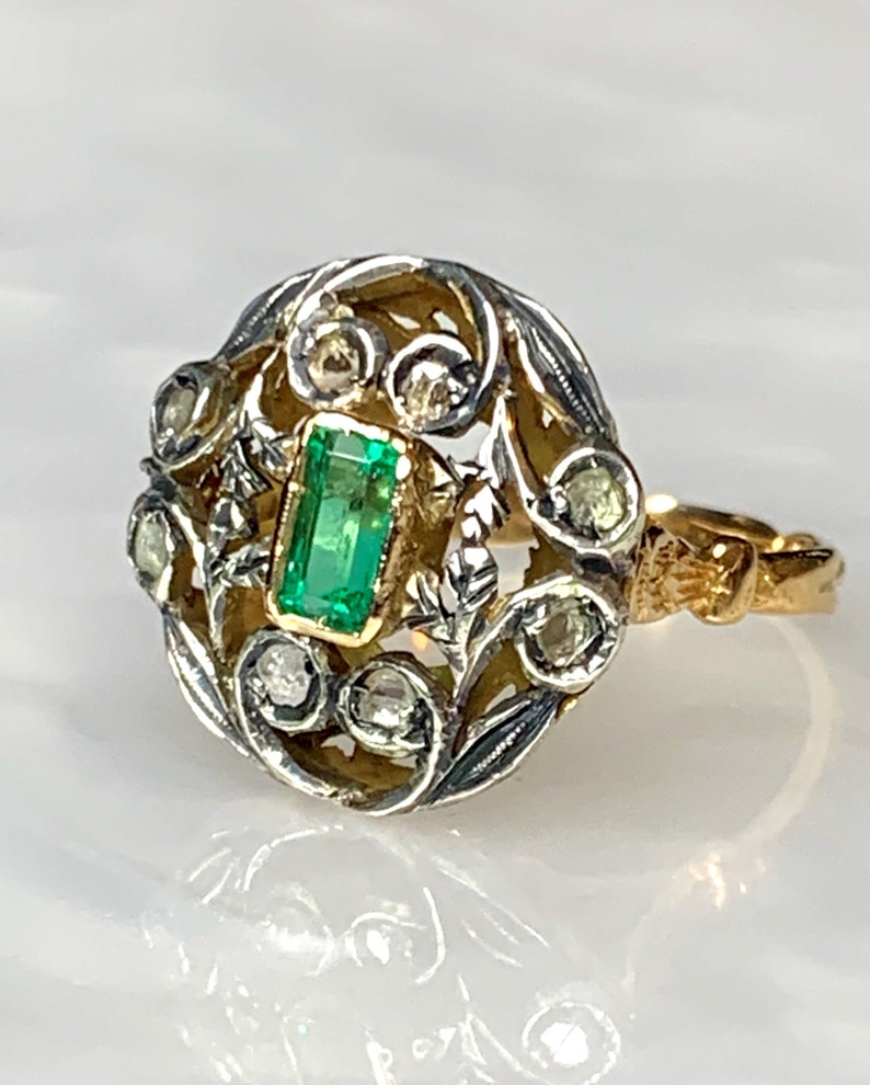 May include: A gold and silver ring with an emerald gemstone surrounded by smaller diamonds. The ring has a delicate, floral design.