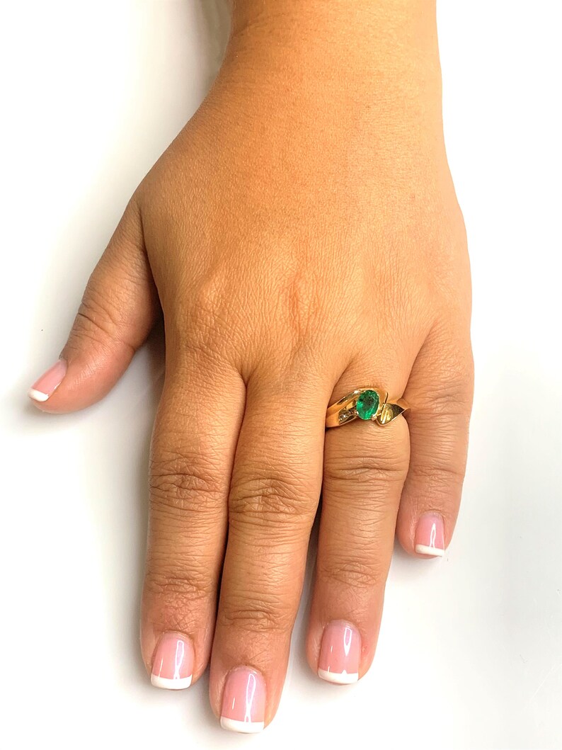 Peut inclure: Une bague en or avec une pierre pr&eacute;cieuse verte sertie dans une bande torsad&eacute;e. La bague est port&eacute;e &agrave; l'annulaire d'une main aux ongles manucur&eacute;s.
