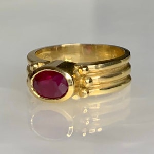 Puede incluir: Un anillo de oro con una piedra preciosa de rubí rojo engastada en un engaste de bisel. La banda tiene tres filas de oro texturizado.