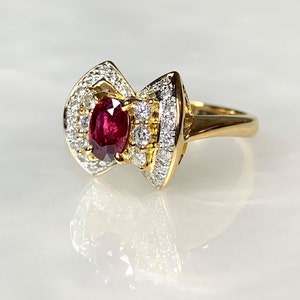 Könnte beinhalten: Ein goldener Ring mit einem großen ovalen Rubin, der von kleineren Diamanten umgeben ist. Der Ring hat ein einzigartiges Design mit zwei geschwungenen Krappen, die sich in der Mitte treffen.
