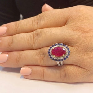 Op de afbeelding: Een gouden ring met een grote ovale robijn omringd door een halo van kleinere diamanten en saffieren.
