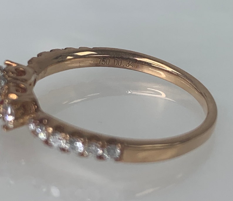 Puede incluir: Un anillo de oro con una piedra preciosa grande rodeada de piedras m&aacute;s peque&ntilde;as. El anillo est&aacute; grabado con "750 DO.34".