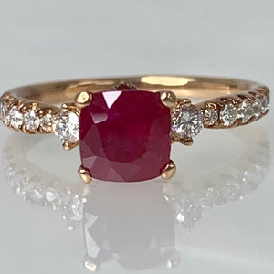 Puede incluir: Un anillo de oro con una gran piedra de rub&iacute; roja cuadrada rodeada de diamantes m&aacute;s peque&ntilde;os. El anillo tiene una banda de diamantes.