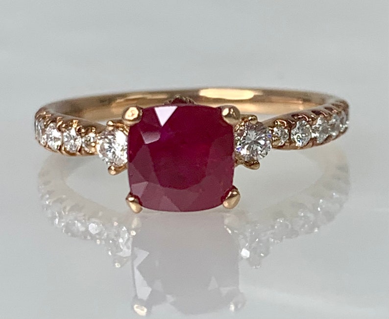 Puede incluir: Un anillo de oro con una gran piedra de rub&iacute; roja cuadrada rodeada de diamantes m&aacute;s peque&ntilde;os. El anillo tiene una banda de diamantes.
