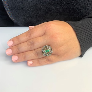 May include: A silver ring with a green emerald gemstone surrounded by smaller diamonds set in a delicate filigree design.