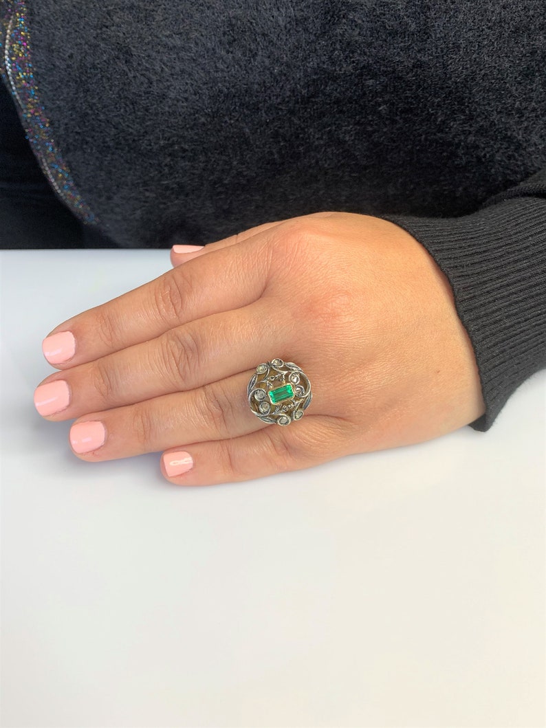 May include: A silver ring with a green emerald gemstone surrounded by smaller diamonds set in a delicate filigree design.