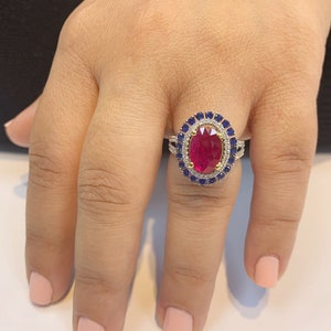 Op de afbeelding: Een gouden ring met een grote ovale robijn, omringd door een halo van kleinere diamanten en saffieren.
