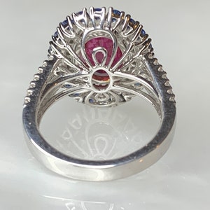 Op de afbeelding: Een zilveren ring met een grote ovale roze edelsteen, omringd door een halo van kleinere blauwe edelstenen en diamanten.