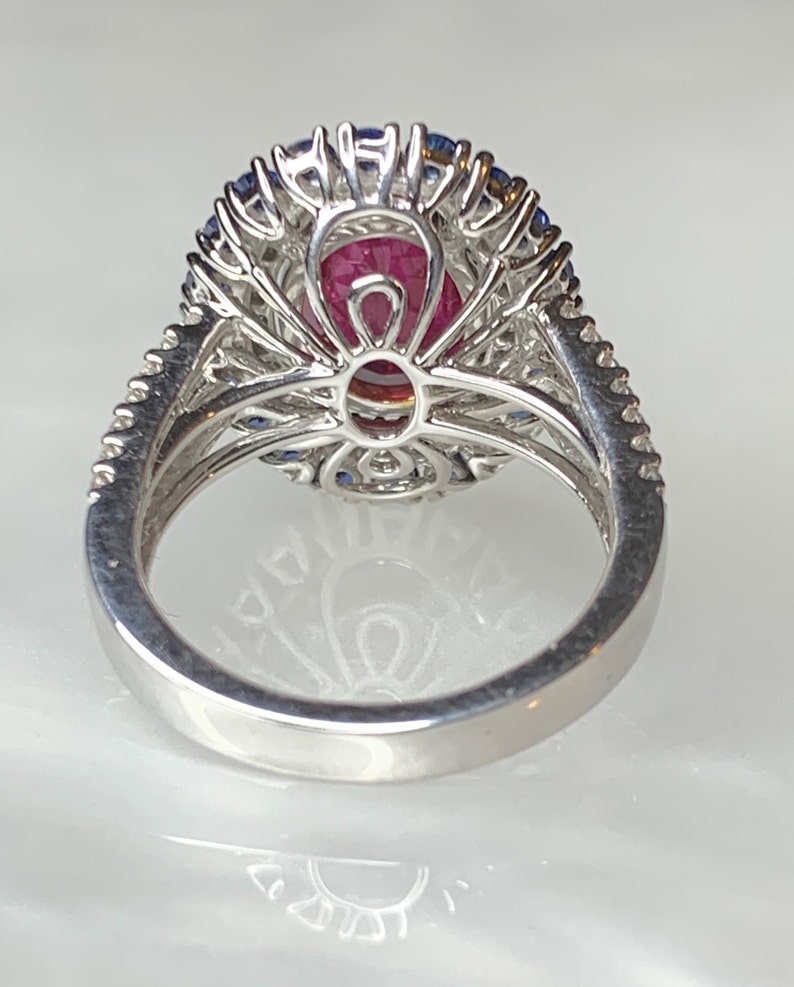 Op de afbeelding: Een zilveren ring met een grote ovale roze edelsteen, omringd door een halo van kleinere blauwe edelstenen en diamanten.