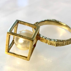 May include: A gold ring with a textured band and a square cage holding a pearl.