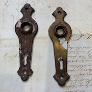 May include: Two antique metal door handle backplates. One is a dark, rusty brown, and the other is a lighter brass color. Both have a keyhole and a decorative top with a hole for a screw. The backplates show signs of wear.