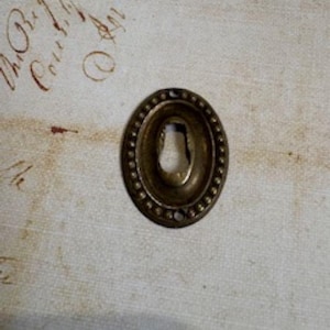 May include: Oval-shaped antique brass keyhole cover with a beaded border. The keyhole is centered within the oval, and the brass has a slightly aged appearance. The background is a textured, off-white surface with faded script.