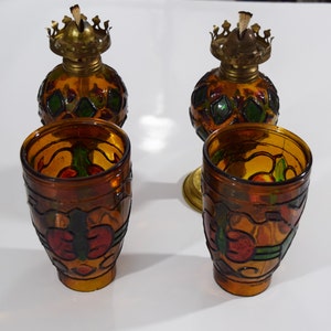 May include: Set of four amber glass oil lamps with stained glass designs. The lamps have a round body with a metal top and a wick. The designs feature geometric shapes and floral motifs in shades of red, green, and yellow.