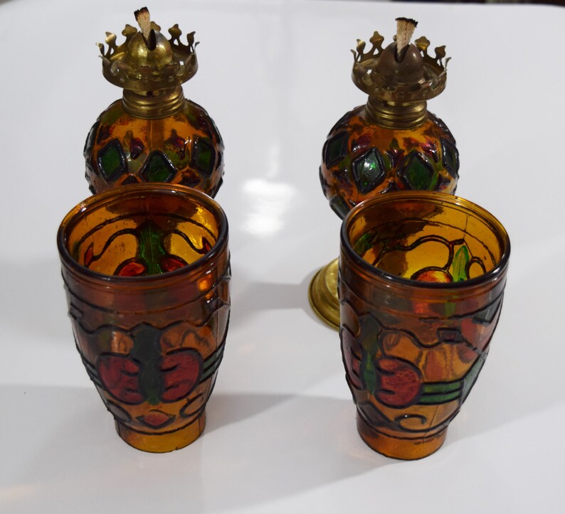 May include: Set of four amber glass oil lamps with stained glass designs. The lamps have a round body with a metal top and a wick. The designs feature geometric shapes and floral motifs in shades of red, green, and yellow.