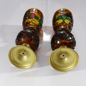 May include: A pair of amber glass candle holders with stained glass designs in shades of red, green, and yellow. The candle holders have a round base with a gold finish.