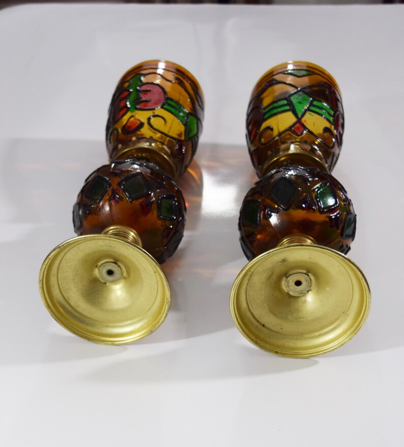 May include: A pair of amber glass candle holders with stained glass designs in shades of red, green, and yellow. The candle holders have a round base with a gold finish.