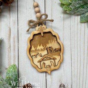 May include: A wooden Christmas ornament with a cutout design depicting the nativity scene. The ornament is brown and features a star, a city skyline, and the year 2021.
