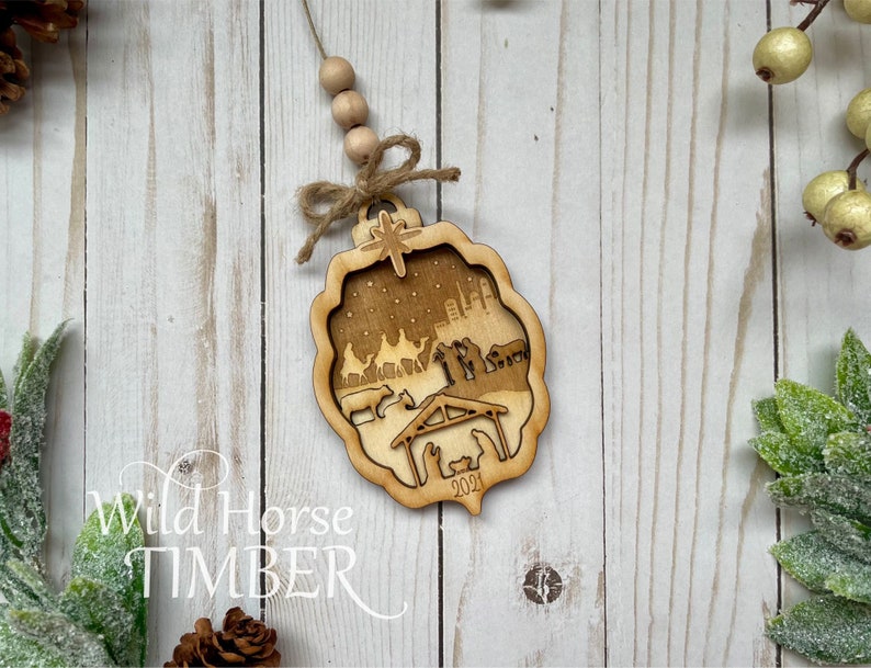 May include: A wooden Christmas ornament featuring a nativity scene with a star on top. The ornament is laser cut and has a rustic brown finish. The text "Wild Horse Timber" and "2021" are visible on the ornament.