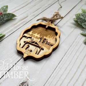 May include: A wooden Christmas ornament with a laser-cut design of the nativity scene. The ornament is brown and features a star, a stable, and figures of Mary, Joseph, and baby Jesus. The year 2021 is inscribed on the ornament.