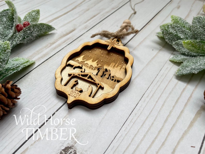 May include: A wooden Christmas ornament with a laser-cut design of the nativity scene. The ornament is brown and features a star, a stable, and figures of Mary, Joseph, and baby Jesus. The year 2021 is inscribed on the ornament.