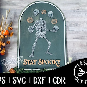 Puede incluir: Un letrero de pizarra verde con un esqueleto haciendo malabares con calabazas. El letrero dice "Stay Spooky".
