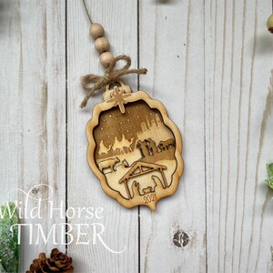 May include: A wooden Christmas ornament featuring a nativity scene with a star on top. The ornament is laser cut and has a rustic brown finish. The text "Wild Horse Timber" and "2021" are visible on the ornament.