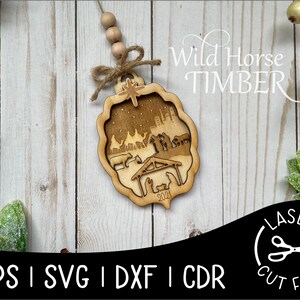 May include: A wooden Christmas ornament featuring a laser-cut nativity scene with the text "2021". The ornament is hanging from a string with wooden beads. The background is a white woodgrain surface. The text "Wild Horse Timber" is in the upper right corner of the image. The text "EPS | SVG | DXF | CDR" is in the lower left corner of the image. The text "LASER CUT FILE" is in the lower right corner of the image.