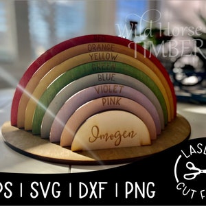 Puede incluir: Juguete de arcoíris de madera con siete arcos en tonos de naranja, amarillo, verde, azul, violeta, rosa y madera natural. Los arcos están apilados uno encima del otro y el arco inferior tiene el nombre "Imogen" grabado en él. El juguete está sobre una base de madera.