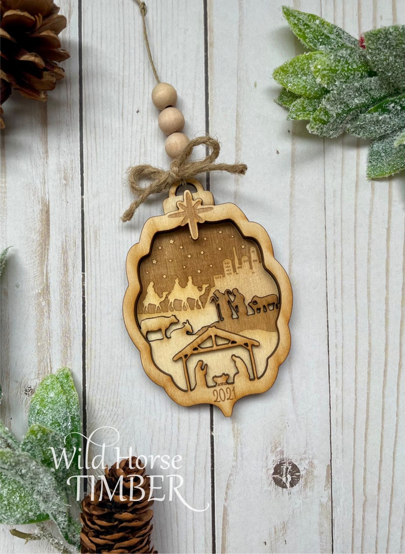 May include: A wooden Christmas ornament with a cutout design depicting the nativity scene. The ornament is brown and features a star, a city skyline, and the year 2021.