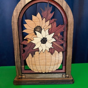 May include: A wooden cutout of a pumpkin with autumn leaves and flowers in a decorative archway frame. The frame is made of dark wood with a brown stain. The pumpkin is made of light wood with a natural finish. The leaves are made of red and brown wood. The flowers are made of white and yellow wood.