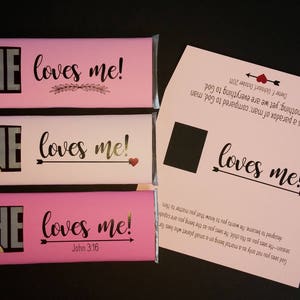 May include: Three pink chocolate bar wrappers with the text "HE loves me!" on each. The top wrapper has a decorative branch design. The middle wrapper has a gold arrow and heart. The bottom wrapper has the text "John 3:16".