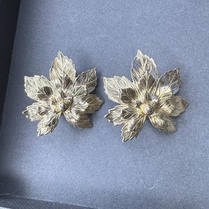 Peut inclure: Une paire de boucles d'oreilles en métal doré en forme de feuilles d'érable superposées. Chaque boucle présente une surface texturée et nervurée avec plusieurs segments de feuilles pointus rayonnant autour d'une petite perle métallique centrale.