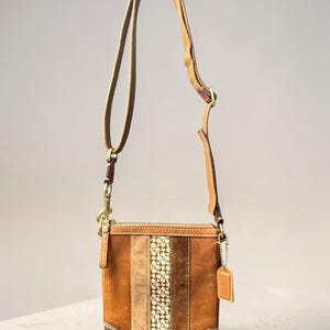 May include: A tan leather crossbody bag featuring a vertical central panel with a repeating logo pattern flanked by suede and leather stripes. The bag includes an adjustable shoulder strap, brass-toned hardware, and a small leather hangtag.