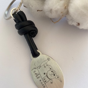 May include: A silver oval keyring with a black leather strap and a silver key ring. The keyring features a hand-drawn illustration of a person with a bird on their head and the text "E.E.E.E. ROPAPA".