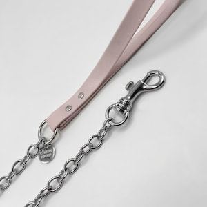 Puede incluir: Una correa de perro de cuero rosa con una cadena plateada y un clip plateado. La correa tiene una pequeña etiqueta plateada con el texto "Urban Pup" en ella.