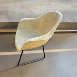 May include: A vintage, off-white molded plastic chair with a curved seat and backrest. The chair has a black metal base with four legs. The chair is designed for comfort and style, suitable for various interior settings.