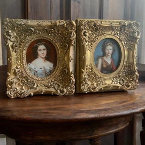 May include: Two antique gold-colored framed portraits. Each frame has an ornate, detailed design. The portraits are oval-shaped, featuring women in period clothing. One portrait has a red background, the other a blue background. The frames sit on a dark wooden table.